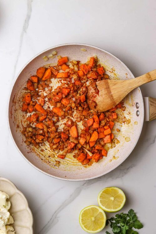 Sautéed vegetables in a white skillet with a wooden spatula, lemon halves, cauliflower, and cilantro on marble surface.
