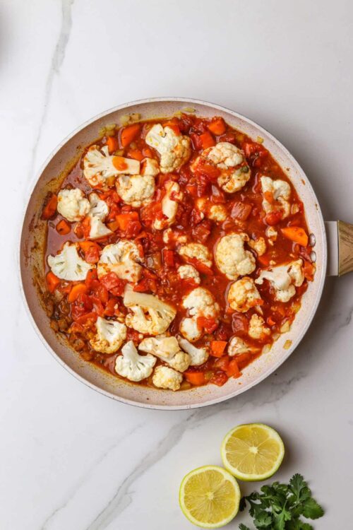 Savory cauliflower stew with diced carrots, tomatoes, and herbs in a skillet on white surface.