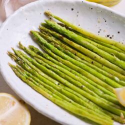 Roasted asparagus on white plate with lemon wedges and olive oil in background.