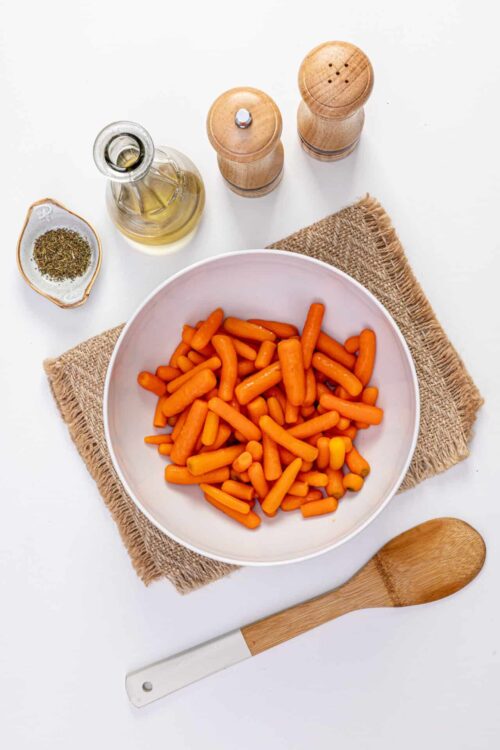 Roasted carrot sticks in a white bowl with olive oil, salt, and pepper shakers on a white background.