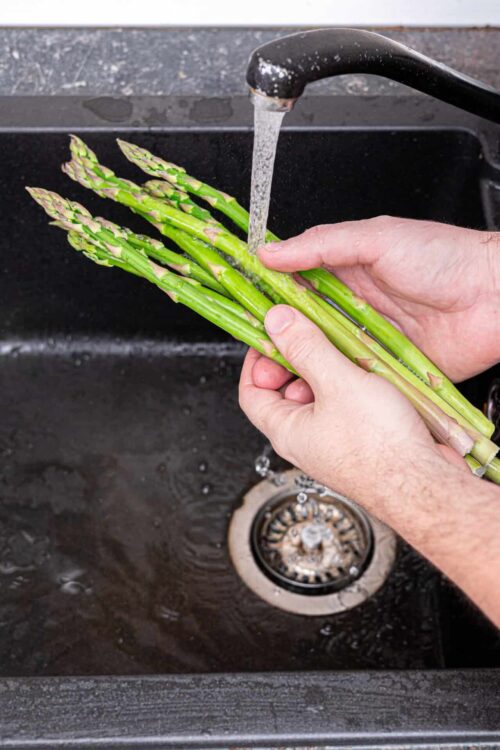 Fresh asparagus being washed under running water in a kitchen sink.