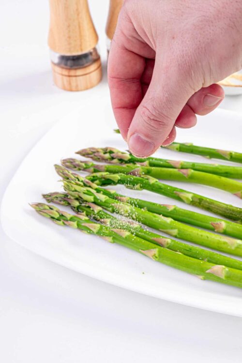 Roasted asparagus being seasoned with salt on a white plate.