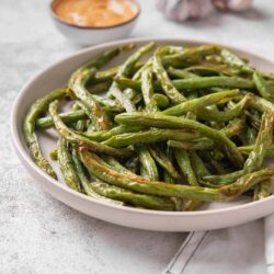 Roasted green beans on white plate with garlic and dipping sauce.