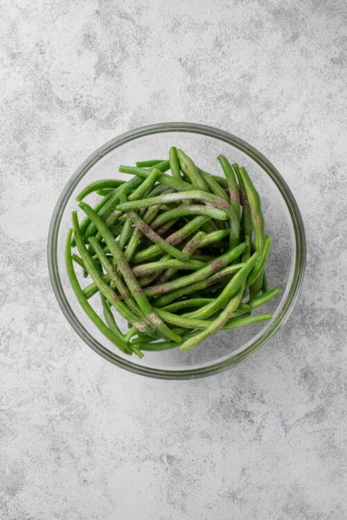 Fresh green beans in a clear glass bowl on textured grey background.