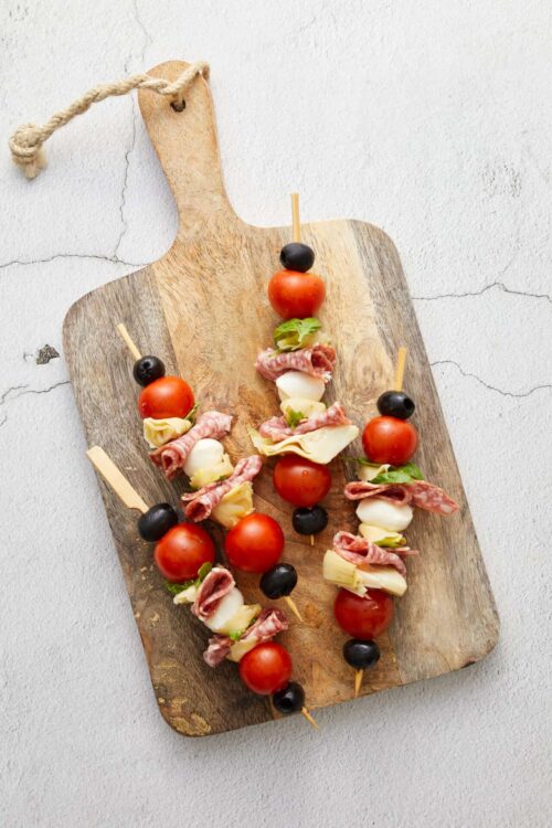 Fresh caprese skewers with cherry tomatoes, mozzarella, basil, prosciutto, and black olives on a rustic wooden serving board.