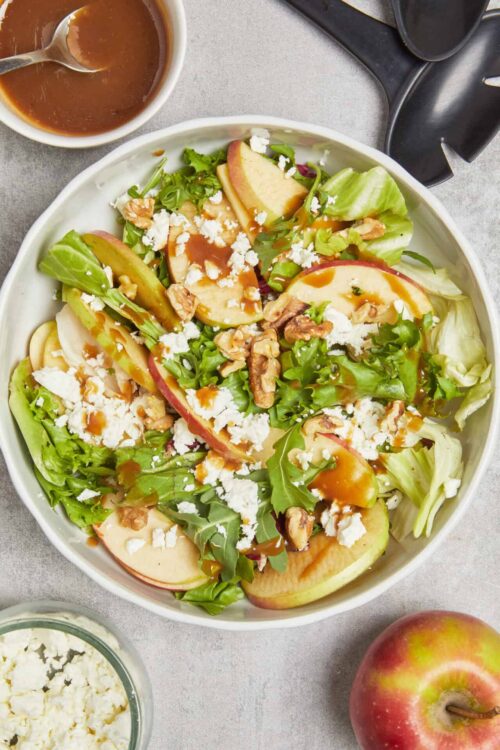 Fresh apple and mixed greens salad with walnuts, feta, balsamic glaze, and vinaigrette on a white bowl.