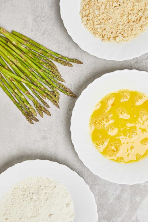 Fresh asparagus, breadcrumb mixture, beaten eggs, and grated cheese on white plates for asparagus casserole.