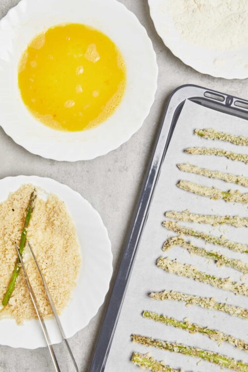 Baked Bree - Preparing breaded asparagus for baking with egg wash, bread crumbs, and parchment-lined baking sheet.