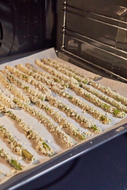 Thin asparagus spears coated in breadcrumbs and Parmesan cheese, baking on a parchment-lined tray.
