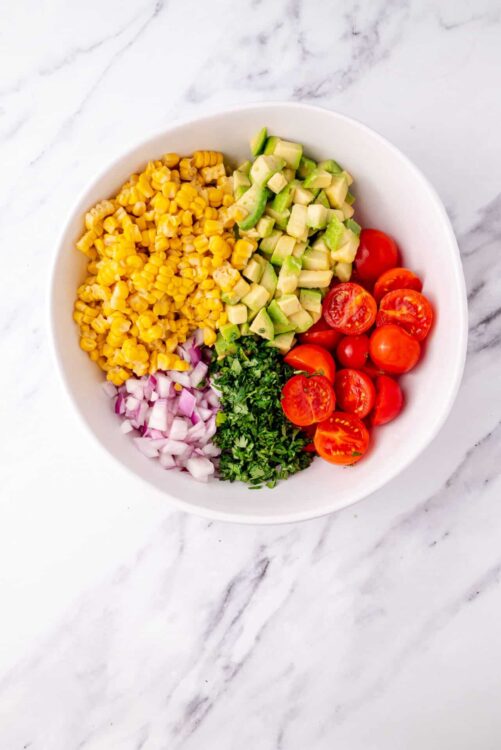Fresh chopped vegetables in a white bowl, including cherry tomatoes, corn, avocado, onion, and cilantro, for healthy eating and meal prep.