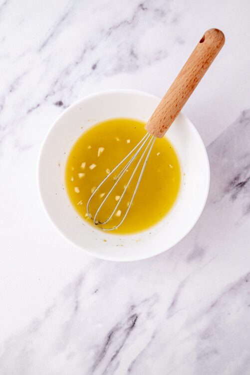 Golden olive oil and minced garlic in a white bowl with a whisk, ready for cooking or salad dressing.