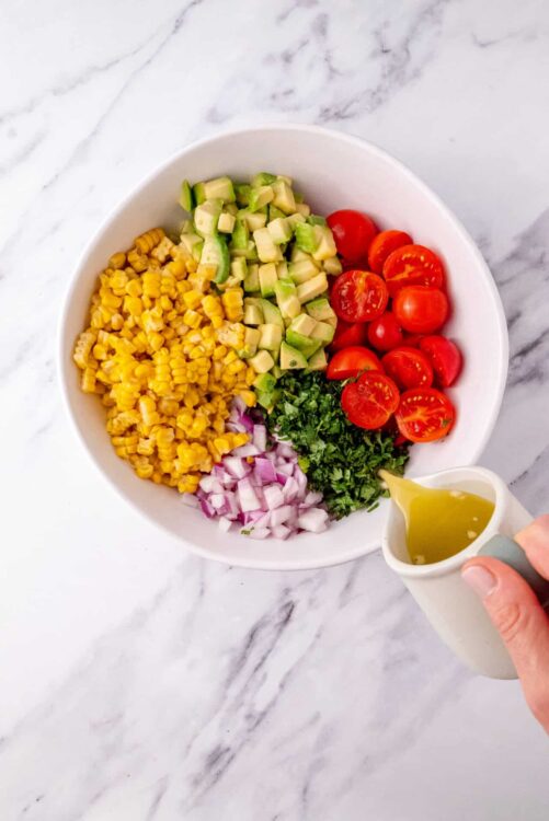 Fresh vegetable salad ingredients in a white bowl with dressing being poured, cherry tomatoes, corn, cucumber, cilantro, red onion, healthy lunch, colorful vegetables.