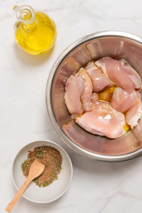 Raw chicken breasts in stainless steel bowl with olive oil, seasonings, and oil bottle on white surface.