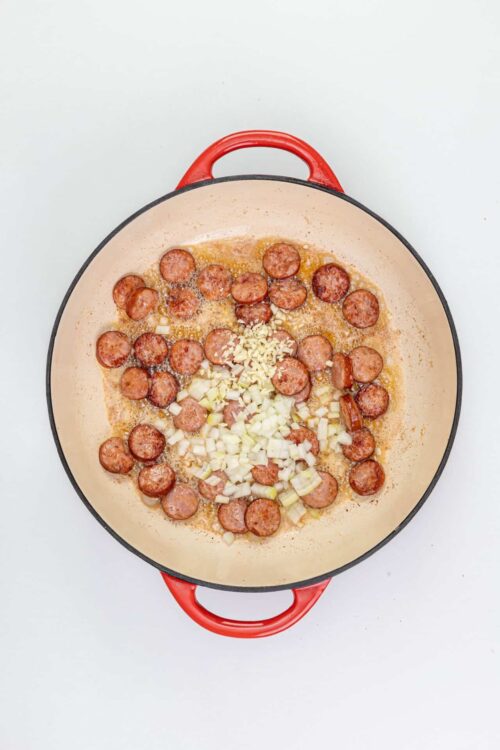 Sautéed sausage slices with chopped onions and garlic in a skillet on a white background.