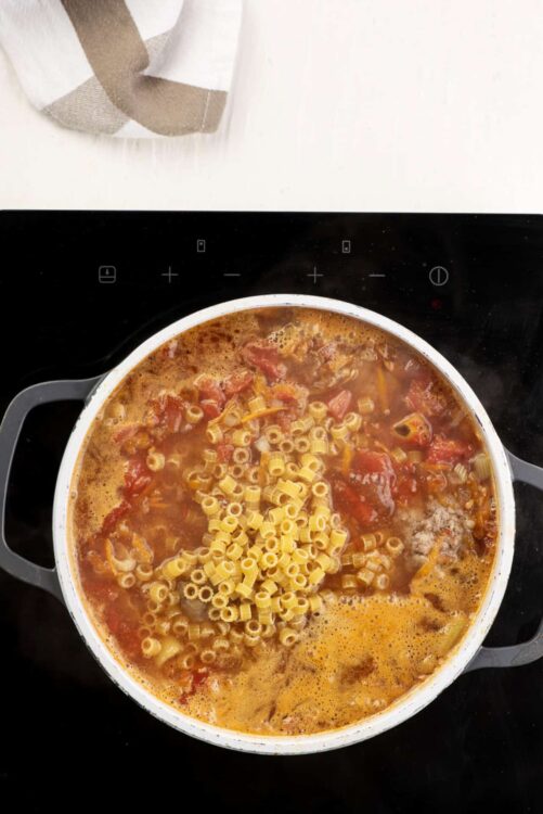 Creamy tomato pasta soup cooking in a white pot on a black stovetop.