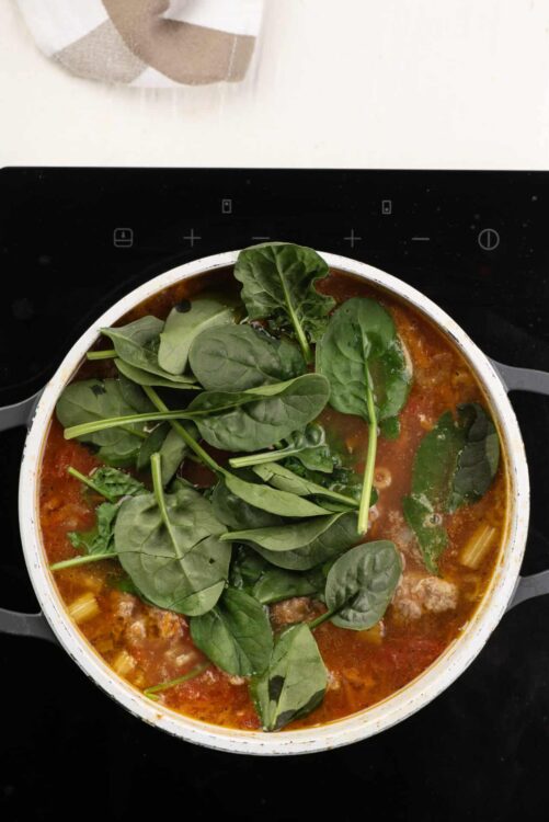 Fresh spinach added to homemade tomato soup in a white pot on a black stovetop.