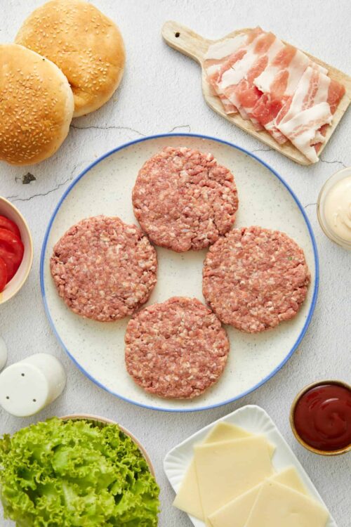Fresh uncooked hamburger patties on a plate with ingredients for burgers around it, including buns, bacon, cheese, lettuce, and condiments.