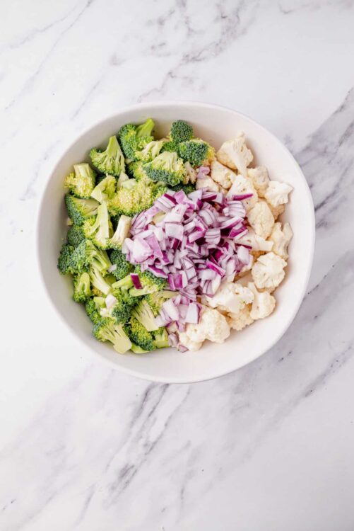 Raw broccoli, cauliflower, and red onions in a white bowl on marble surface.