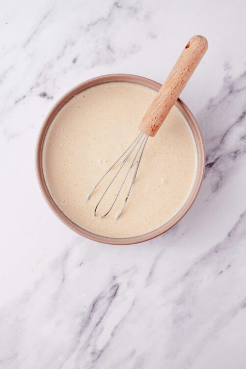 Fluffy homemade pancake batter with whisk in a bowl on marble surface.