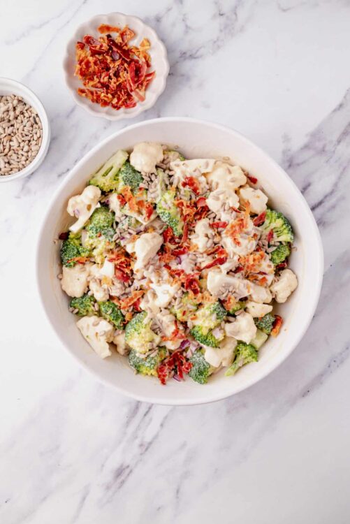 Creamy broccoli cauliflower salad with sunflower seeds, bacon, and red onion on marble surface.