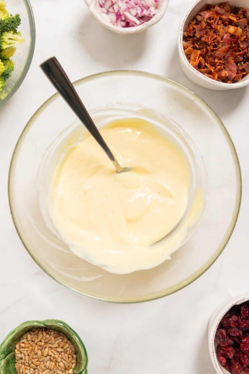 Creamy white cheese sauce in a glass bowl with various chopped vegetables and toppings around it.