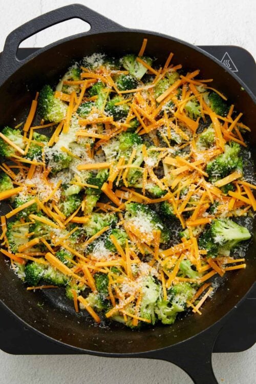 Broccoli and shredded cheddar cheese in a black skillet for baked broccoli cheese casserole.
