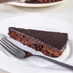 Rich chocolate cake slice with glossy ganache on a white plate.