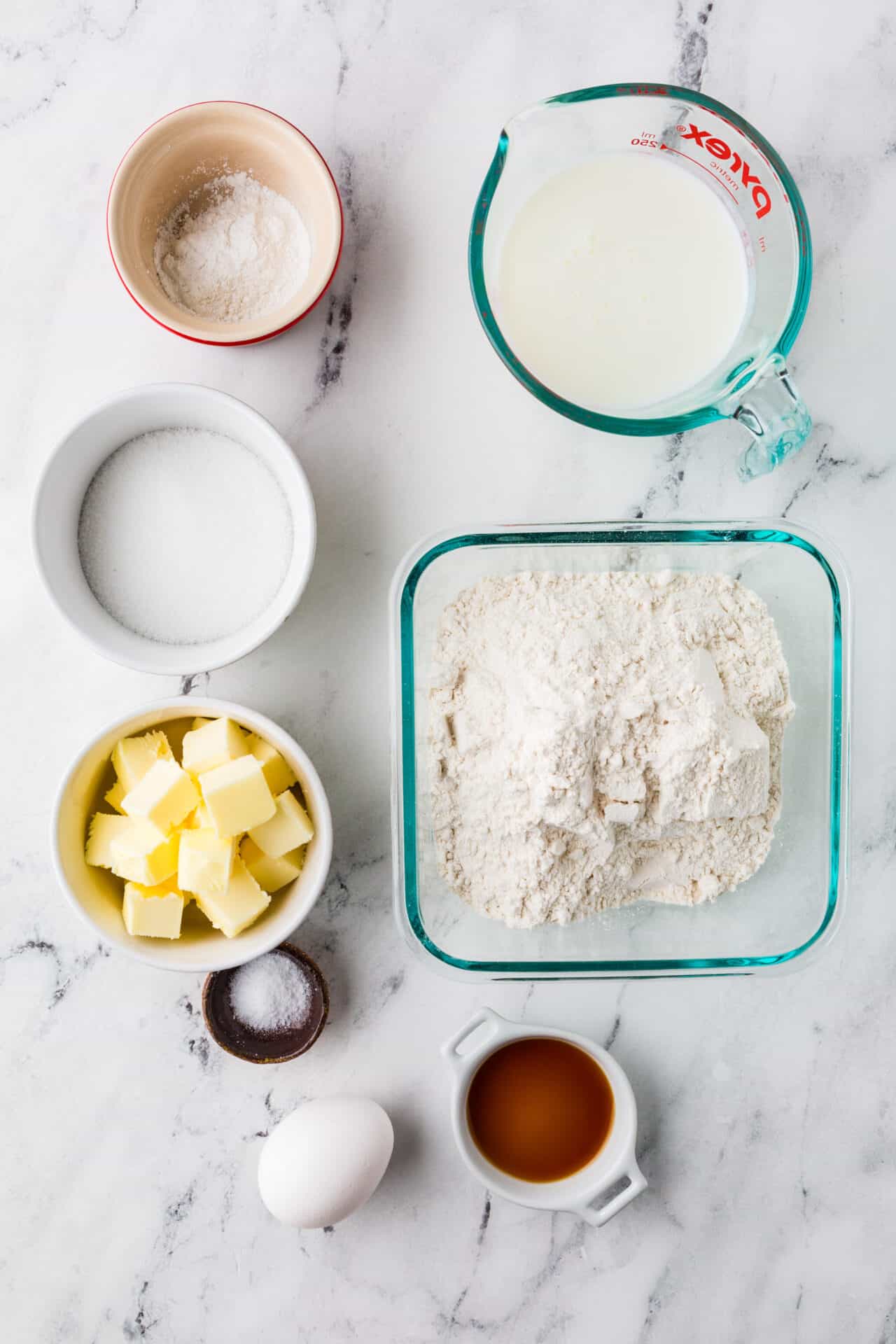 Flour, butter, sugar, milk, vanilla, eggs, salt, and baking ingredients for baked goods on marble surface.