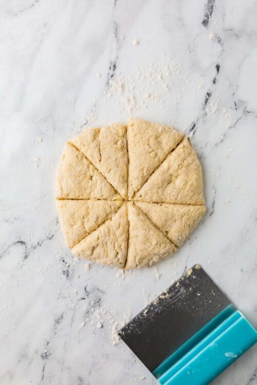 Buttery shortbread dough cut into wedges on marble surface with pastry scraper.