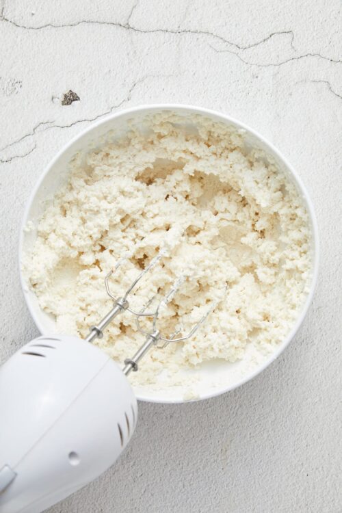 Creamy homemade butter in a white bowl with an electric hand mixer, on a textured white surface.