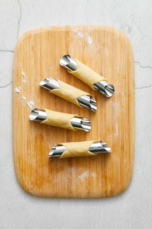 Uncooked pastry dough wrapped around metal cannoli tubes on wooden cutting board.