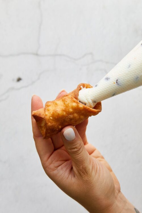 Fried cannoli being filled with creamy ricotta cheese from a piping bag, showcasing classic Italian dessert.