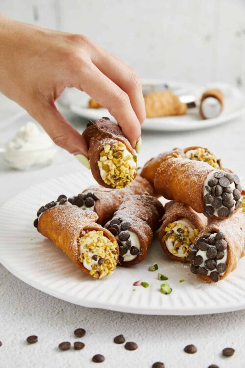 Delicious homemade cannoli on a white plate, filled with cream and topped with chopped pistachios and chocolate chips.