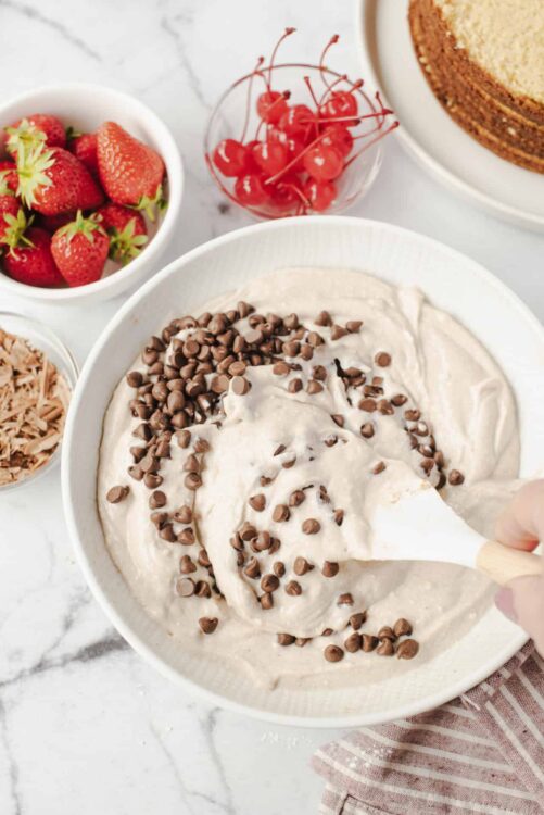 Creamy chocolate chip dip in a white bowl with fresh strawberries and cherries around all on a marble surface.