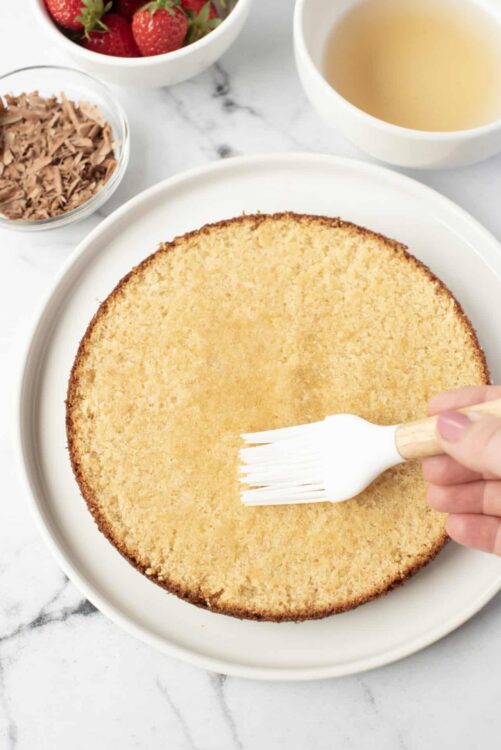 Fluffy yellow vanilla sponge cake on a white plate with a hand brushing it with simple syrup.