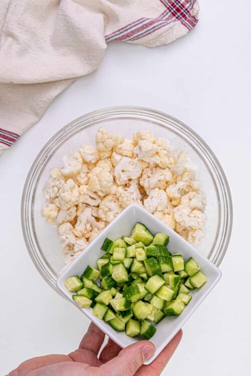 Chopped cauliflower and diced cucumbers in a white bowl on a white surface.
