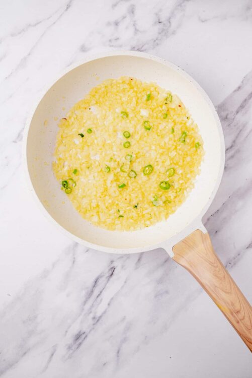 Creamy scrambled eggs with chopped green onions in a white skillet on marble countertop.