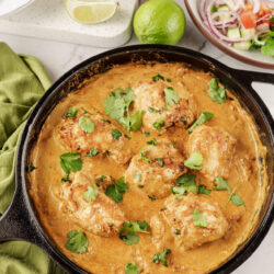 Creamy chicken curry in a black skillet garnished with cilantro and lime wedges on a white marble surface.
