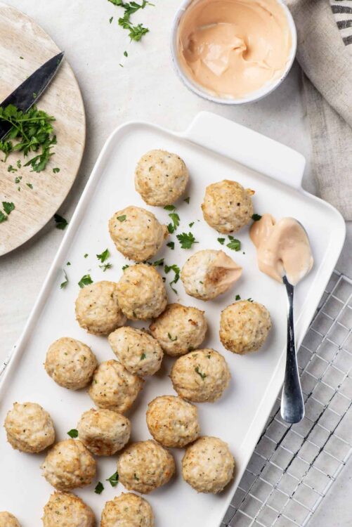 Crispy baked chicken meatballs with parsley on white serving platter, creamy dipping sauce, and fresh herbs.