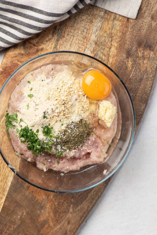 Seasoned raw chicken in a glass bowl with herbs, spices, egg, and butter, ready for cooking or baking.