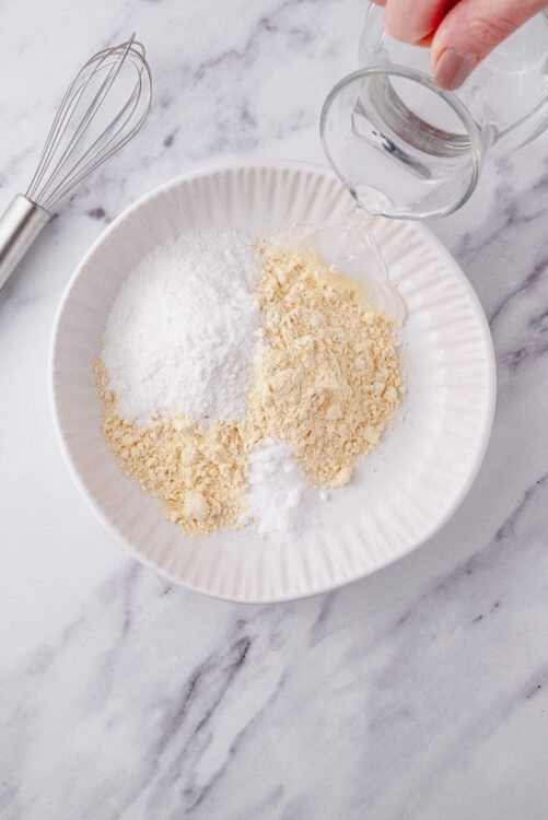 Flour, sugar, and powdered ingredients in a white bowl for baking, with a metal whisk on a marble surface.