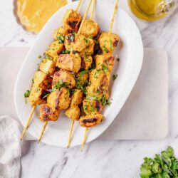 Skewers of grilled chicken tikka with fresh herbs on a white platter with side dipping sauce and olive oil, on a marble surface.
