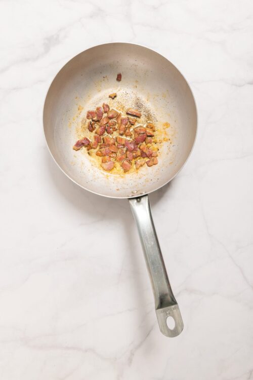 Crispy bacon bits cooking in a skillet on a white marble surface for delicious recipes.
