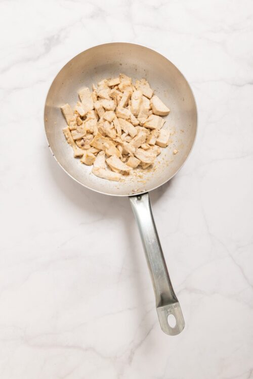 Cooked diced chicken in a stainless steel skillet on a white marble surface.