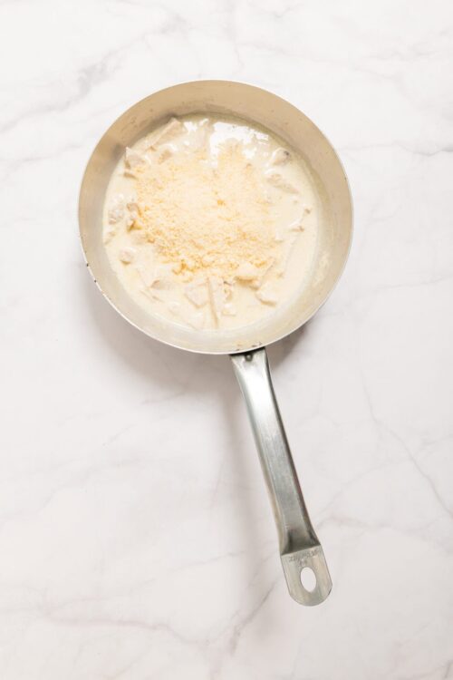 Creamy cheese sauce with shredded cheese in a stainless steel saucepan on white marble background.