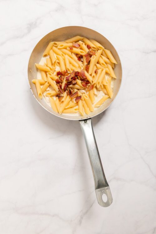 Creamy pasta with bacon bits in a stainless steel skillet, close-up.