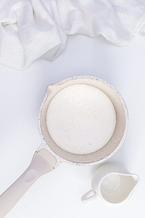 Creamy milk in a small saucepan on a white surface with a white dishtowel and small pitcher.