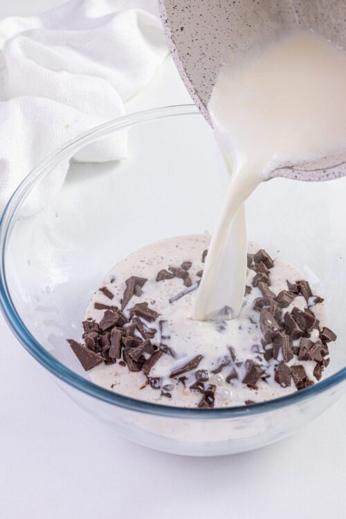 Faintly speckled mixing bowl with pouring cream over chopped chocolate and milk mixture on white background.