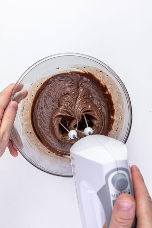 Mixing chocolate frosting with a hand mixer in a glass bowl at Baked Bree.