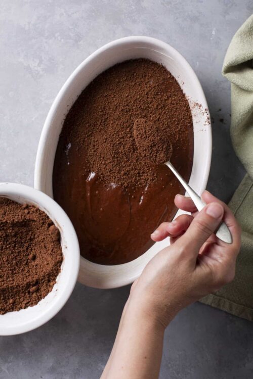 Rich chocolate pudding being topped with cocoa powder in a white baking dish.